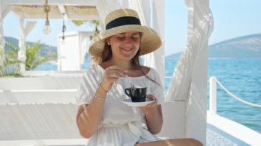 Genç esmer kadın deniz kenarındaki kafede ya da restoranda dinleniyor ve deniz dalgalarına bakarken kahve içiyor. Yaz tatili, tatil, seyahat, yolculuk ve seyahatler kavramı