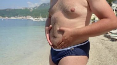 Koca göbekli şişman bir adamın deniz sahilinde yüzmeye gitmeden önce kilolu vücuduna bakması. Sağlık kavramı, zayıflama, diyet, yaz gelmeden kilo verme.