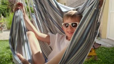 Gülümseyen genç bir çocuk, hamağın bahçedeki nazik salınımında gevşer. Yaz mevsimini, mutlu çocukluk anılarını, boş zamanlarını ve sevgi dolu yaz tatillerini özetler..