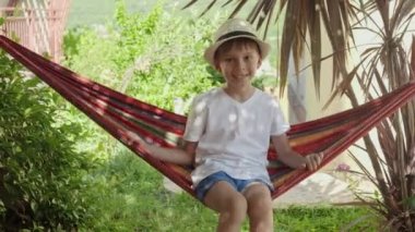 Şapkalı genç bir çocuk bir hamakta oturur ve sallanır, tasasız çocukluk anılarını yakalar ve bir tatilin rahatlığını yakalar..