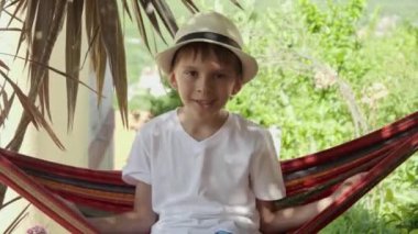 Şapkalı çocuk rahatlıyor, bahçe hamağında sallanıyor, tasasız anıları kucaklıyor ve tatil sükunetini kucaklıyor..