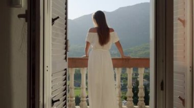 Kabarık elbiseli genç bir kadın, villanın terasında ya da balkonunda duruyor, ve dağ günbatımında keyifli. Yaz zamanı, dağ manzarası, tatil kaçamakları ve seyahat hayalleri..
