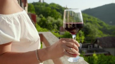 Yakın plan çekimde, elinde kırmızı şarap kadehi olan bir kadın, güneş batarken ve villa balkonundan ya da terastan dağ manzaralı bir yerde..