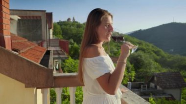 Genç bir kadın, zarif giyinmiş, balkonda ya da villa terasında bir bardak kırmızı şarap içiyor, nefes kesen dağ günbatımının arka planında. Seyahat ruhu, yaz ve tatil mutluluğu..
