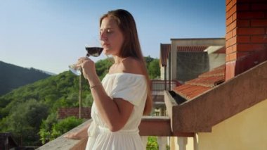 Genç bir kadın, balkonda ya da villa terasında kırmızı şarap yudumlarken dağların ardında büyüleyici günbatımını seyrediyor. Seyahat, yaz tatili ve tatillerin neşesi..