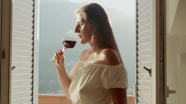 Villa terasında duran bir kadın bardaktan kırmızı şarap yudumlarken, manzaralı dağlar büyüleyici bir zemin görevi görüyor..
