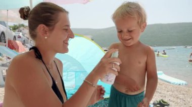 Genç bir anne güneşli bir günde deniz kıyısında dinlenirken bebeğine güneş kremi sürüyor. Sağlık sigortası kavramı, yaz tatili, çocuklarla tatil..