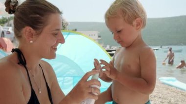 Genç anne, güneşli bir deniz sahilinde dinlenirken bebeğinin hassas cildine dikkatlice güneş kremi sürüyor. Çocuklarla yaz tatillerinde sağlık hizmetlerinin önemi.