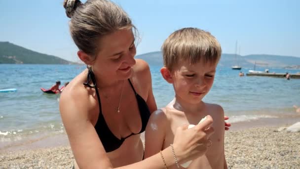 家族の休日の喜び 若い母親は愛情を持って日焼け止めスプレーを息子の穏やかな肌に日焼け止めのビーチに適用します サマータイムの医療 — ストック動画