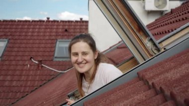 Mutlu gülümseyen ve gülen bir kadın kırmızı kiremitli çatının altındaki evinin açık tavan arasından dışarı bakıyor..