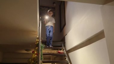 Noel arifesinde Noel Baba 'yı bekleyen heyecanlı küçük çocuk elinde fenerle evin merdivenlerinden aşağı iniyor. Çocuklar kış tatilini kutluyor