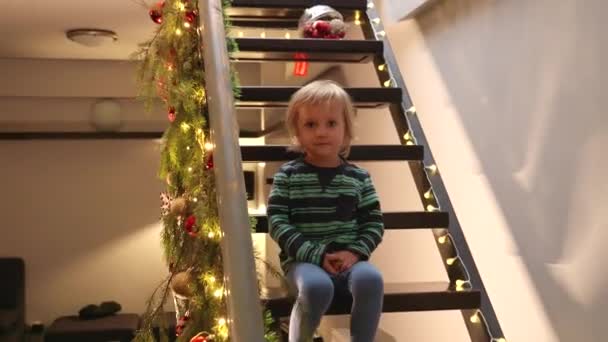 家の装飾された木製の階段に座って クリスマスや新年を待っているかわいい笑顔の幼い少年 冬休みを祝う子供たち — ストック動画