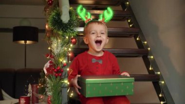 Noel Baba 'nın hediye kutusunu elinde tutan ve evdeki tahta merdivenlerde otururken kameraya gülümseyen mutlu çocuk. Kış tatilinde aile kutlamaları