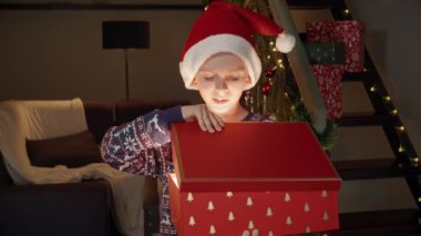 Noel Baba şapkalı heyecanlı küçük çocuk Noel arifesinde hediyesiyle birlikte sihirli, parıldayan Noel hediye kutusunu açıyor. Kış tatilinde aile kutlamaları.