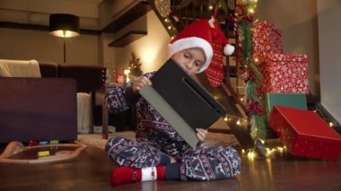 Pijamalı, Noel Baba şapkalı, hediye ve hediyelerin yanında oturup tablet bilgisayarında oyun oynayan mutlu çocuk. Kış tatili, kutlamalar ve parti.