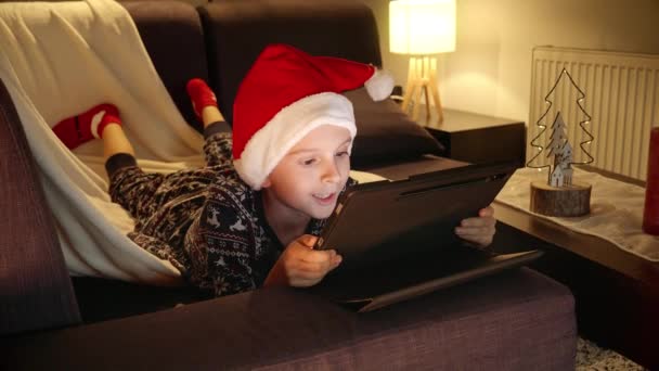Szczęśliwy Uśmiechnięty Chłopiec Piżamie Kapeluszu Mikołaja Leżącego Sofie Korzystającego Tabletu — Wideo stockowe