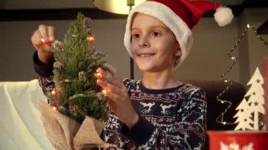 Noel için evi dekore eden küçük şirin bir çocuğun portresi ve küçük bir Noel ağacına ışık konması. Kış tatili, kutlamalar ve parti.