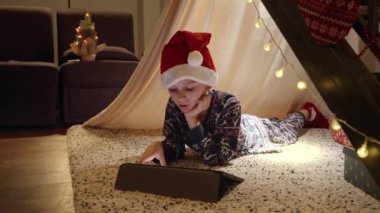 Noel Baba şapkalı küçük çocuk çadırda uzanıyor ve Noel arifesinde tablet bilgisayardan video izliyor. Kış tatili, kutlamalar ve parti.