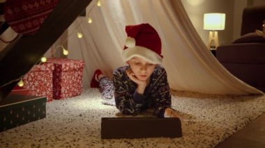 Pijamalı ve Noel Baba şapkalı küçük çocuk çadırda yatıp tablet bilgisayar kullanarak Noel 'i kutluyor. Kış tatili, kutlamalar ve parti.