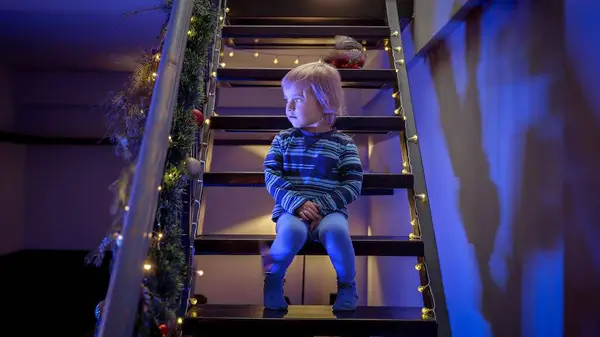 Little boy sitting on stairs decorated for Christmas or New Year and looking at police lights and siren. Concept of Crime on winter holidays, theft, burglar in house and protection.