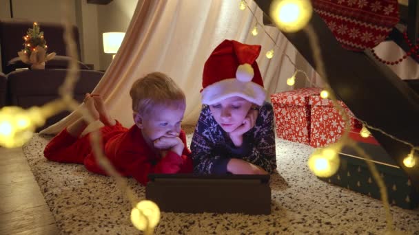 クリスマスの夜にタブレットのコンピュータで漫画を見ている2人の小さな少年は 照明やガーランドが装飾された家で 冬休み お祝い パーティー — ストック動画