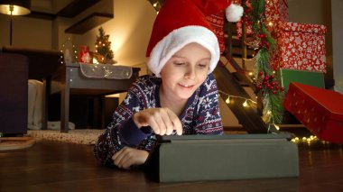 Noel için süslenmiş ve tablet bilgisayarla oynayan mutlu çocuk evde yerde yatıyor. Kış tatili, kutlamalar ve parti.