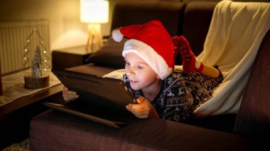 Noel Baba şapkalı küçük çocuğun portresi Noel 'de tablet bilgisayarla oturma odasındaki kanepede yatıyor..