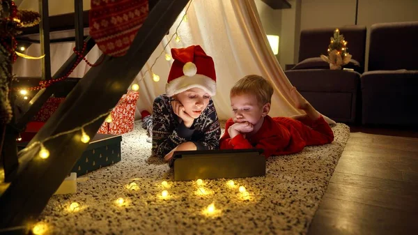 两个身穿睡衣庆祝圣诞节的兄弟躺在地板上 在帐篷里看平板电脑上的视频 庆祝活动和聚会 — 图库照片