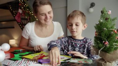 Mutlu gülümseyen anne oğluna bakarak geleneksel Noel kartı ve el yapımı süslemeler yapıyor..