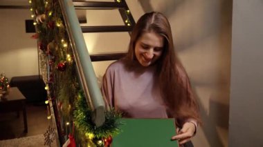 Genç, mutlu, güler yüzlü bir kadın ahşap merdivenlerde oturuyor ve Noel hediyesini bir kutuda açıyor. Kış tatili, yeni yılı kutlamak.