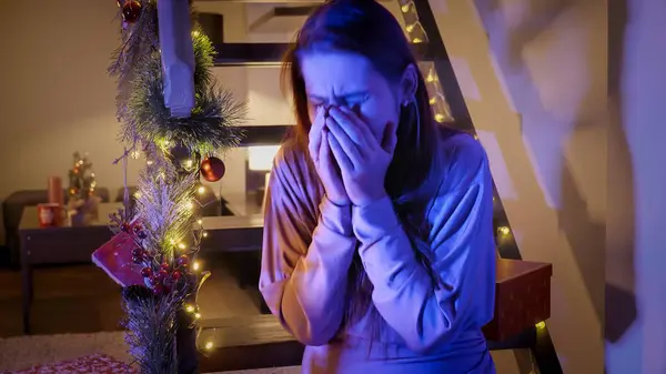 Young crying woman sitting in living room decorated for Christmas or New Year and looking at police lights and sirens. Crime, ambulance, injury, problems with law and theft on winter holidays and celebrations