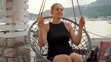 Gün batımında deniz kıyısındaki barda sallanan ve gevşeyen ve sallanan gülümseyen genç kadın. Tatil, yaz tatili, tatil, deniz yolculuğu ve seyahat kavramı.