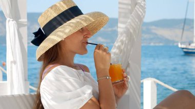 Büyük şapkalı, uzun elbiseli, samanla kokteyl içen yaz tatilinde çardakta dinlenen zarif genç bir kadın..