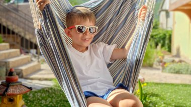 Güneş gözlüklü neşeli bir çocuk bir bahçede sallanıyor ve bir hamakta dinleniyor, yazın ruhunu yayıyor, mutlu çocukluk, rahatlama ve yaz tatillerinin neşesi..