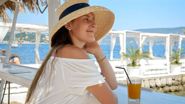 Portrait of elegant woman in straw hat relaxing in beach club bar and looking at the sea view.