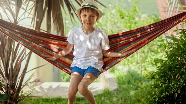 戴帽子的小男孩坐在花园的吊床上打滚 捕捉着无忧无虑的童年回忆和度假的放松 免版税图库图片