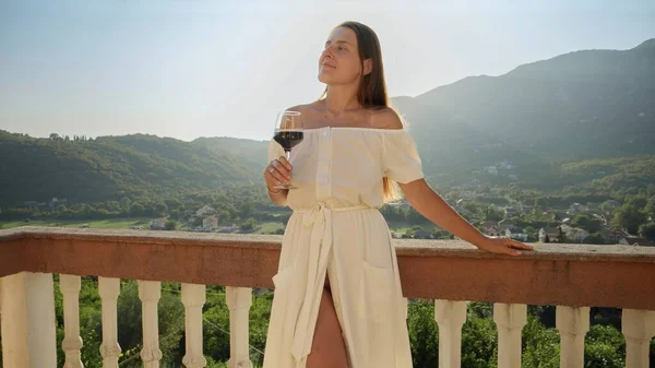 Young woman in a flowing dress relishes a glass of red wine on the balcony or villa terrace, while taking in the magic of a mountain sunset. Travel, summertime, and holiday tranquility.
