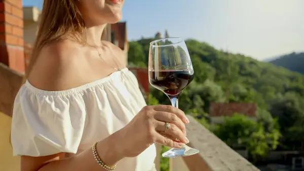若いブルネットの女性のクローズアップは 息をのむような夕日とヴィラのバルコニーやテラスからの山の景色に対して赤ワインのガラスを保持しています 夏時間 休日のリラックス ロイヤリティフリーのストック写真