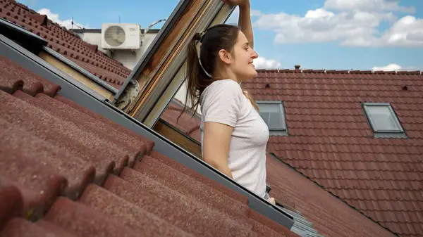 女人笑着从阁楼的窗户往外看 在红色瓷砖屋顶下 呼吸着新鲜空气 图库图片