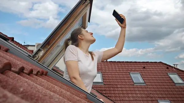 若い女性は 開いた屋根裏窓から外を見ながら スマートフォンでGpsまたは5Gの信号問題に遭遇し 接続を復元することを決定 ストック写真