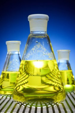 Laboratuvar tezgahında duran sarı sıvılı cam deney şişelerini kapatın ve arkadan ışıklandırın..