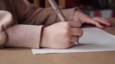 Yedi yaşında bir kız, Masada Evinde oturuyor ve bir kağıt parçasına yazı yazıyor. Yakın çekim. 4K Çözünürlüğü.