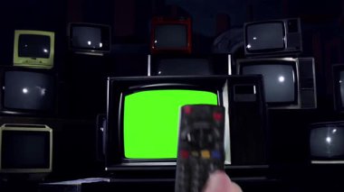 Eski Televizyonlarla çevrili yeşil renkli anahtar ekranlı eski bir televizyonu uzaktan kumandayla açmak, Dolly In -% 100 Gerçek ve yapay zeka içermeyen - 4K