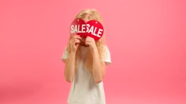 Happy cheerful teenage girl in white T-shirt dances and smiles, closes her eyes with two advertising banners with inscription Sale on pink studio background. Black Friday, big discount mock-up banner