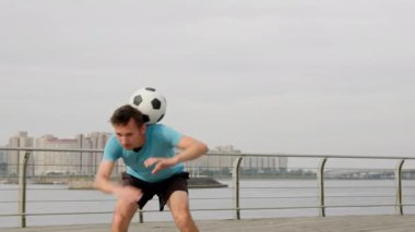 Man professional soccer player performing tricks and doing complex of exercises with football ball on waterfront in city. Footballer is training outdoors in freestyle. Sports skills concept.