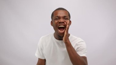 Portrait of positive african american man wearing white t-shirt is shouting with smile holding hands near mouth on white background. Enthusiastic emotions, attracting attention, advertizing.