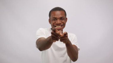 Positive young smiling happy african american man in white t-shirt is pointing fingers to you looking at camera on white background. He is choosing you. Good energy and vibes concept.