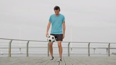 Man footballer juggling kicking bouncing a ball on waterfront in city. Soccer player practicing training outdoors juggling ball improving sport skills in slow motion. Sports football skills concept.