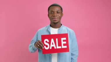 African-American in denim shirt raises banner with inscription sale, rejoices, shouts with emotion, advertises big discount and sale with low prices with place for layout on pink studio background