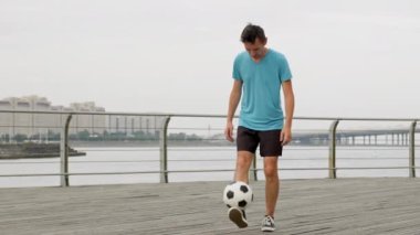 Man soccer player kicking bouncing a ball performing tricks on waterfront. Footballer practicing training outdoors juggling ball improving sport skills in slow motion. Sports football skills concept.
